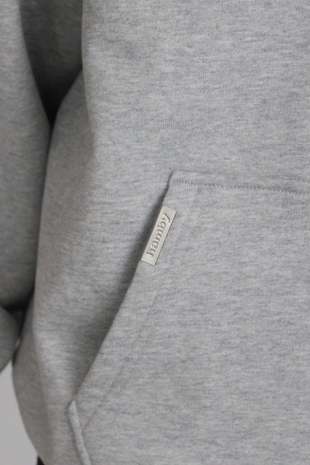 Close up of the hamby metal label in a silver matte finish that is attached to the kangaroo pocket of the hoodie. The metal label states ‘hamby’.