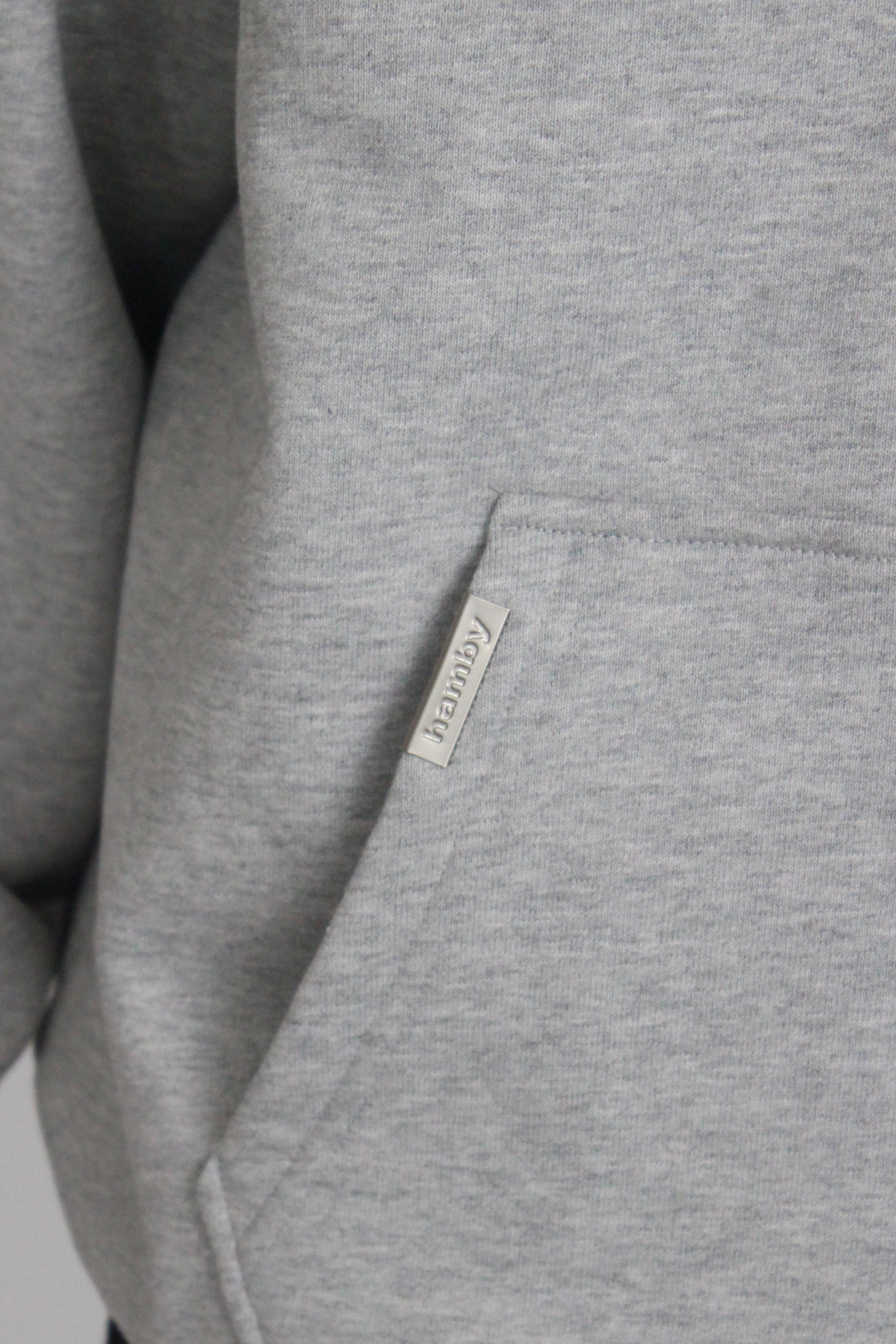 Close up of the hamby metal label in a silver matte finish that is attached to the kangaroo pocket of the hoodie. The metal label states ‘hamby’.