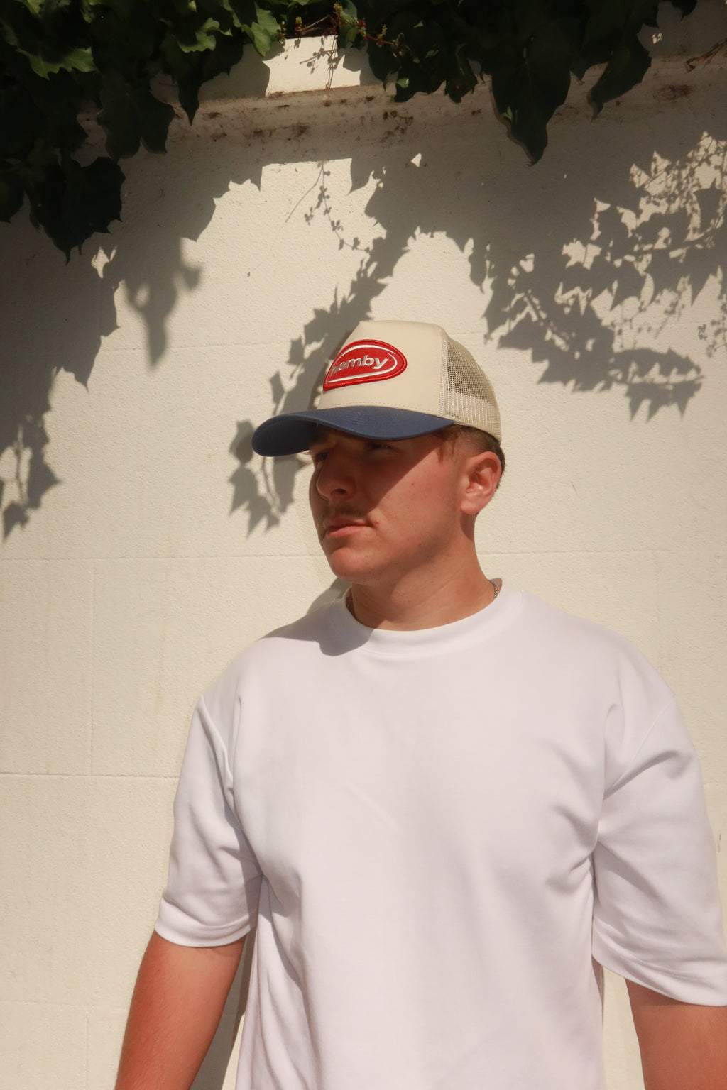 Model wearing hamby's lucky trucker cap. Deep red hamby logo patch, on an off white structured crown with a vintage blue visor.