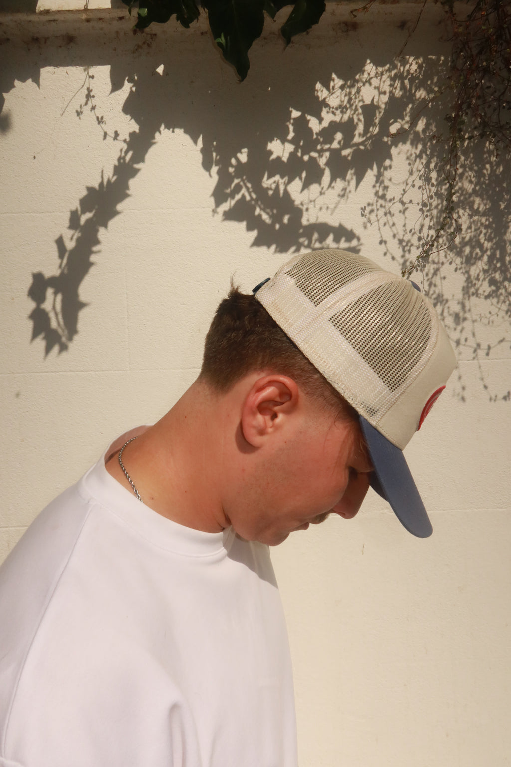 Model wearing hamby's lucky trucker cap. Deep red hamby logo patch, on an off white structured crown with a vintage blue visor. Lucky trucker has a blue snapback closure and red hamby label that's attached to it.