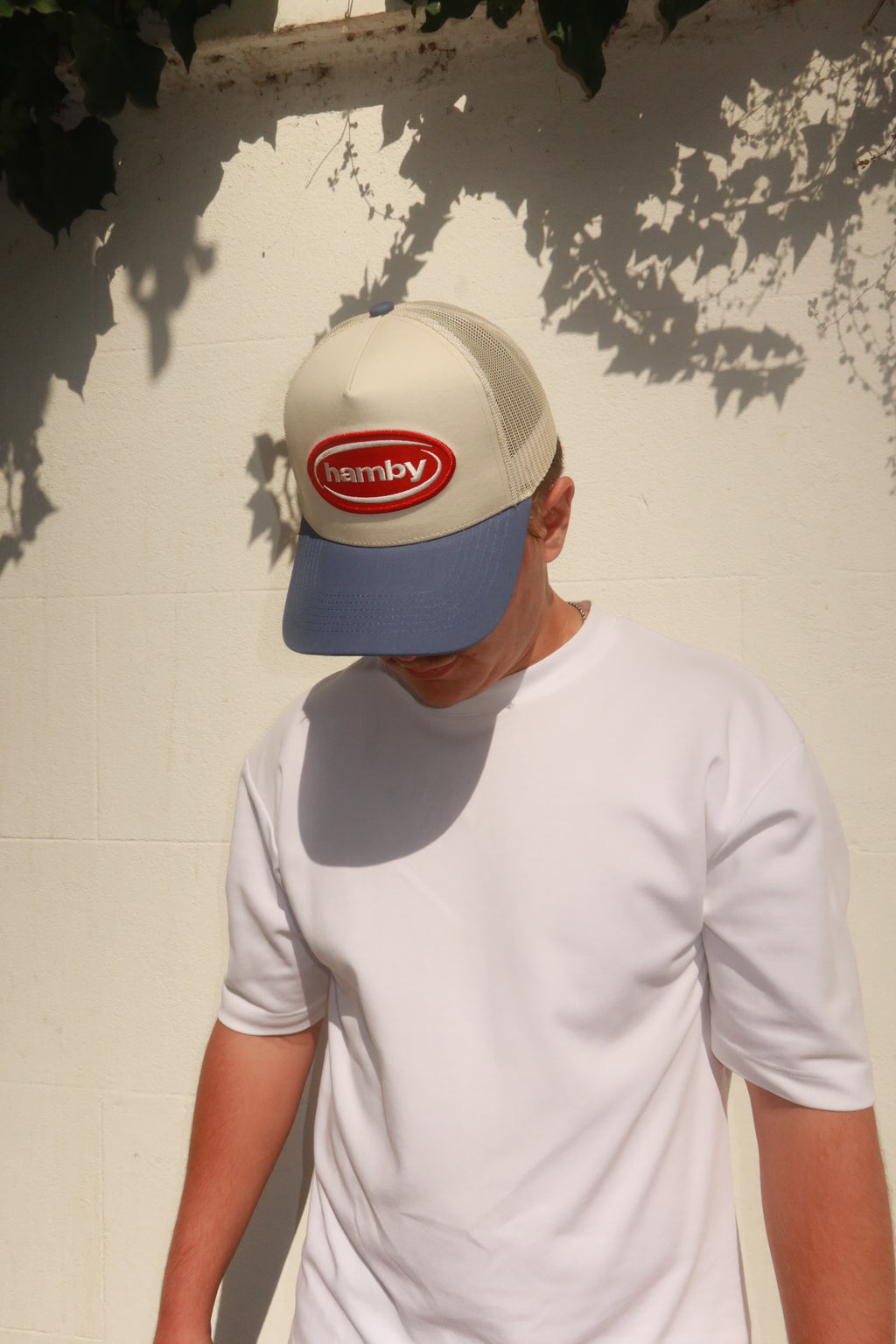 Model wearing hamby's lucky trucker cap. Deep red hamby logo patch, on an off white structured crown with a vintage blue visor.