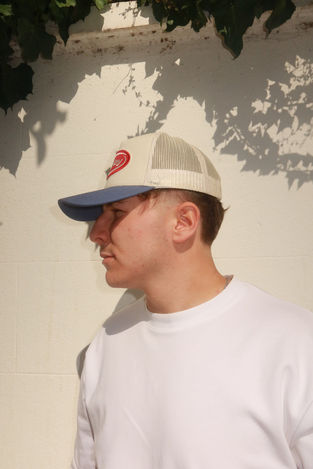 Model wearing hamby's lucky trucker cap. Deep red hamby logo patch, on an off white structured crown with a vintage blue visor.
