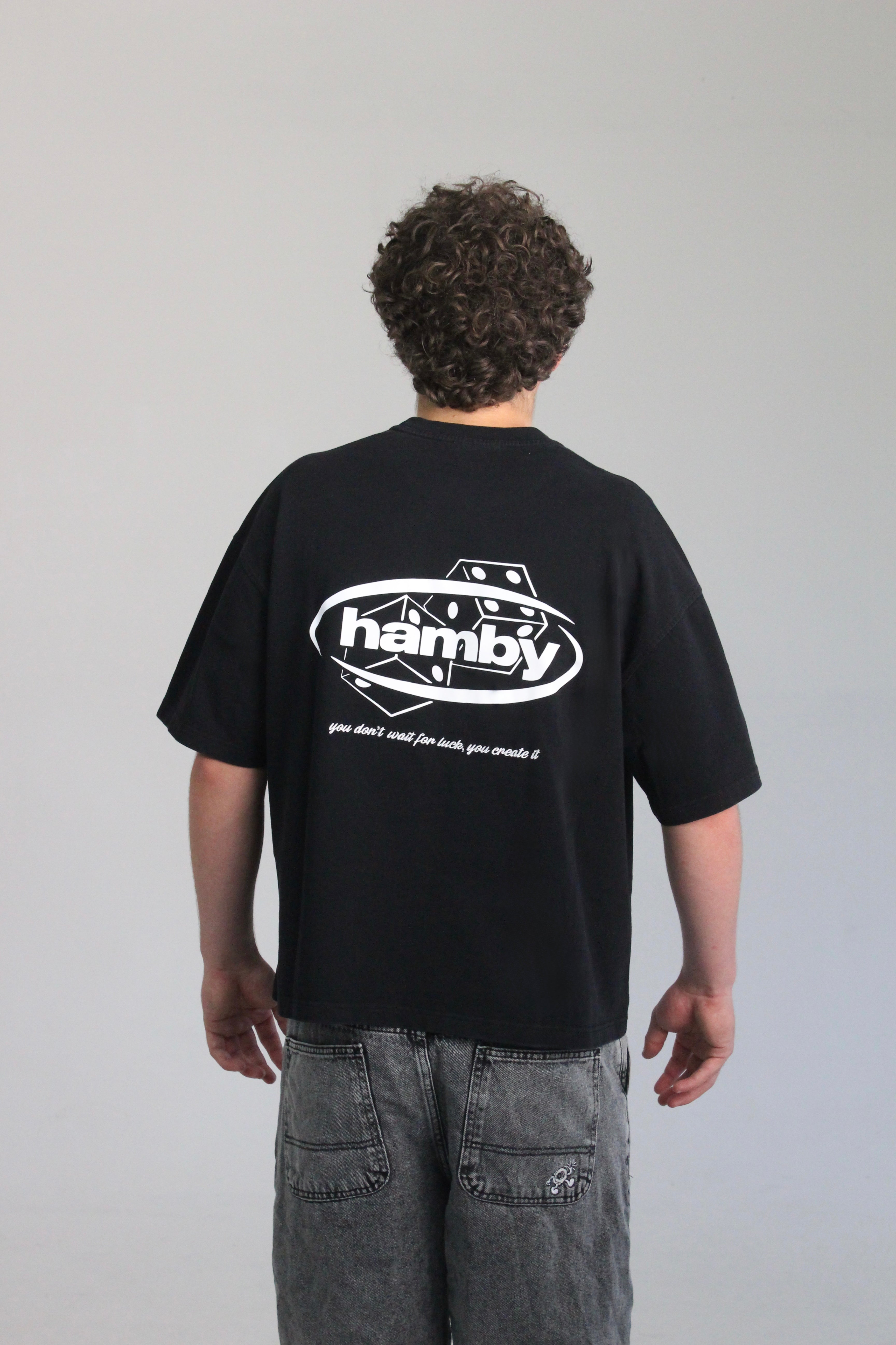 Model wearing the roll the dice tee by hamby in black. The hamby logo screen printed on the back with the signature dice. Then an embroidered quote underneath to complete the design on the heavyweight t-shirt. Quote- ‘you don’t wait for luck, you create it’