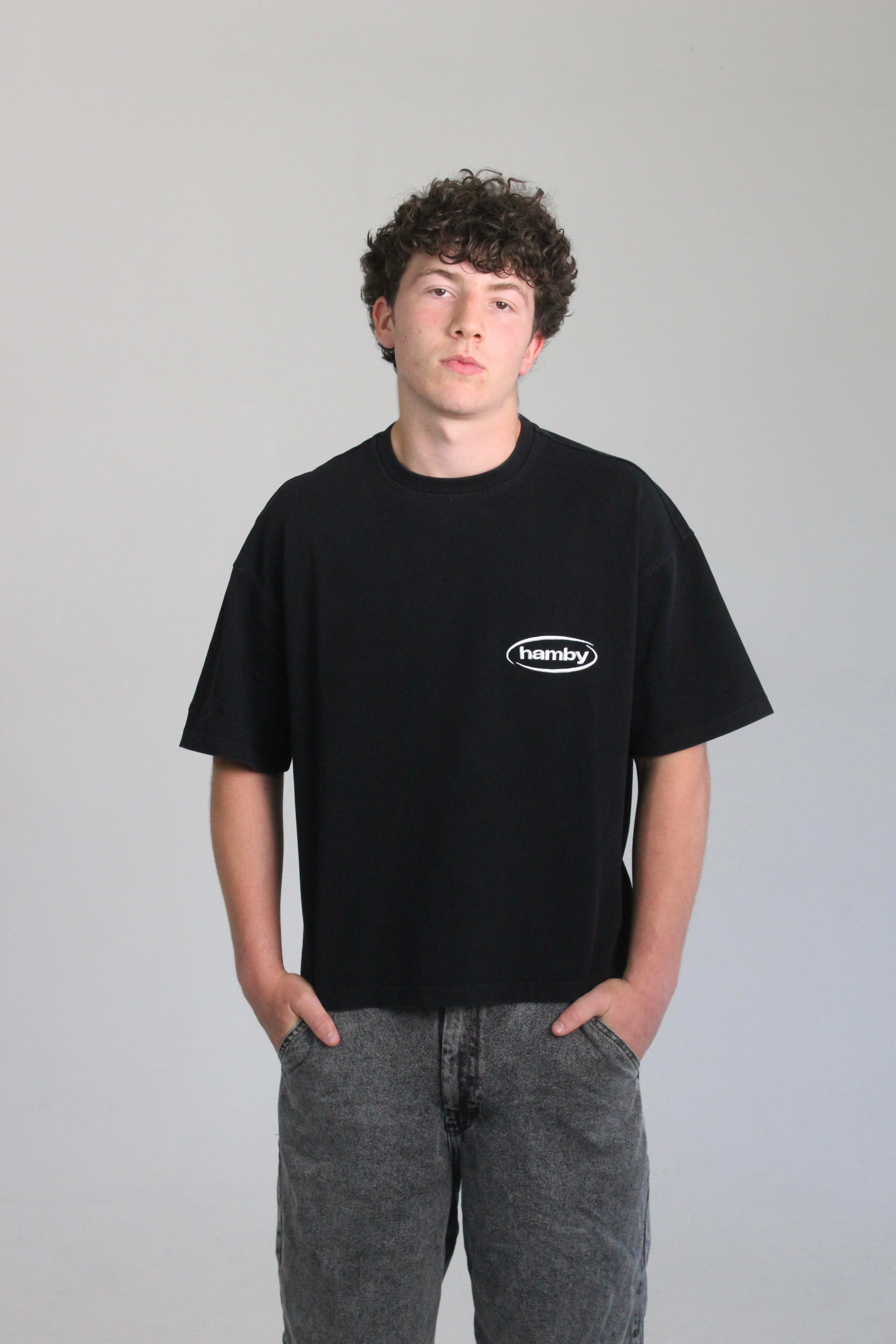 Model wearing boxy oversize hamby black t-shirt with hamby logo embroidered on the front left chest in white. T-shirt is 100% cotton, 240gsm. 