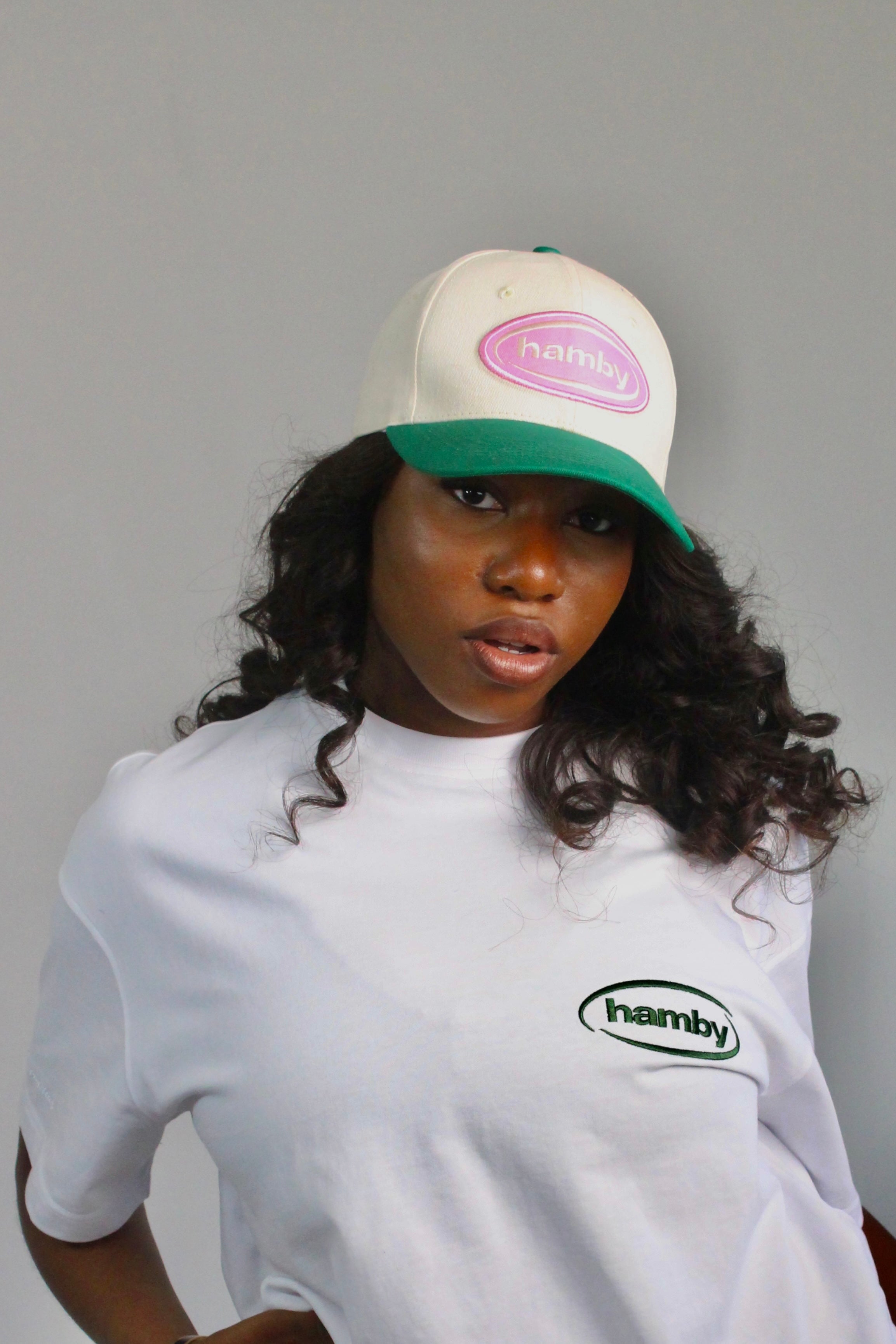 Model wearing a hamby SnapBack baseball cap. Cream cap with pink logo patch and green visor. ‘Create your own luck’ embroidered onto the left panel. 