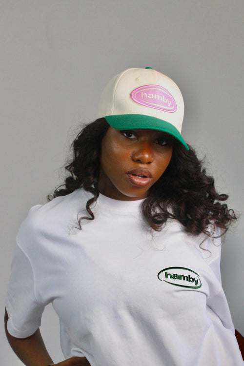 Model wearing a hamby SnapBack baseball cap. Cream cap with pink logo patch and green visor. ‘Create your own luck’ embroidered onto the left panel. 