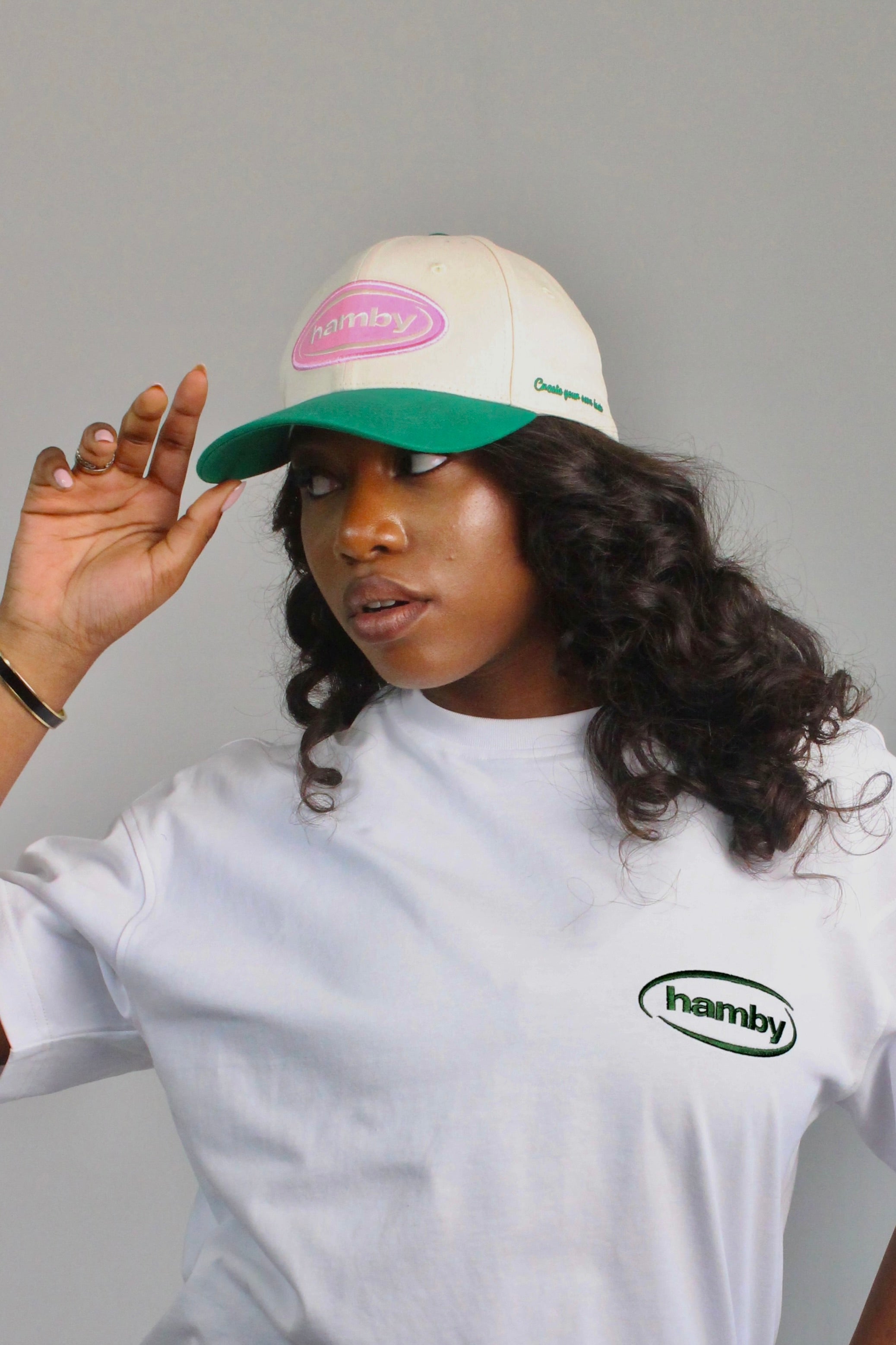 Model wearing a hamby SnapBack baseball cap. Cream cap with pink hamby logo patch and green visor. ‘Create your own luck’ embroidered onto the left panel. 