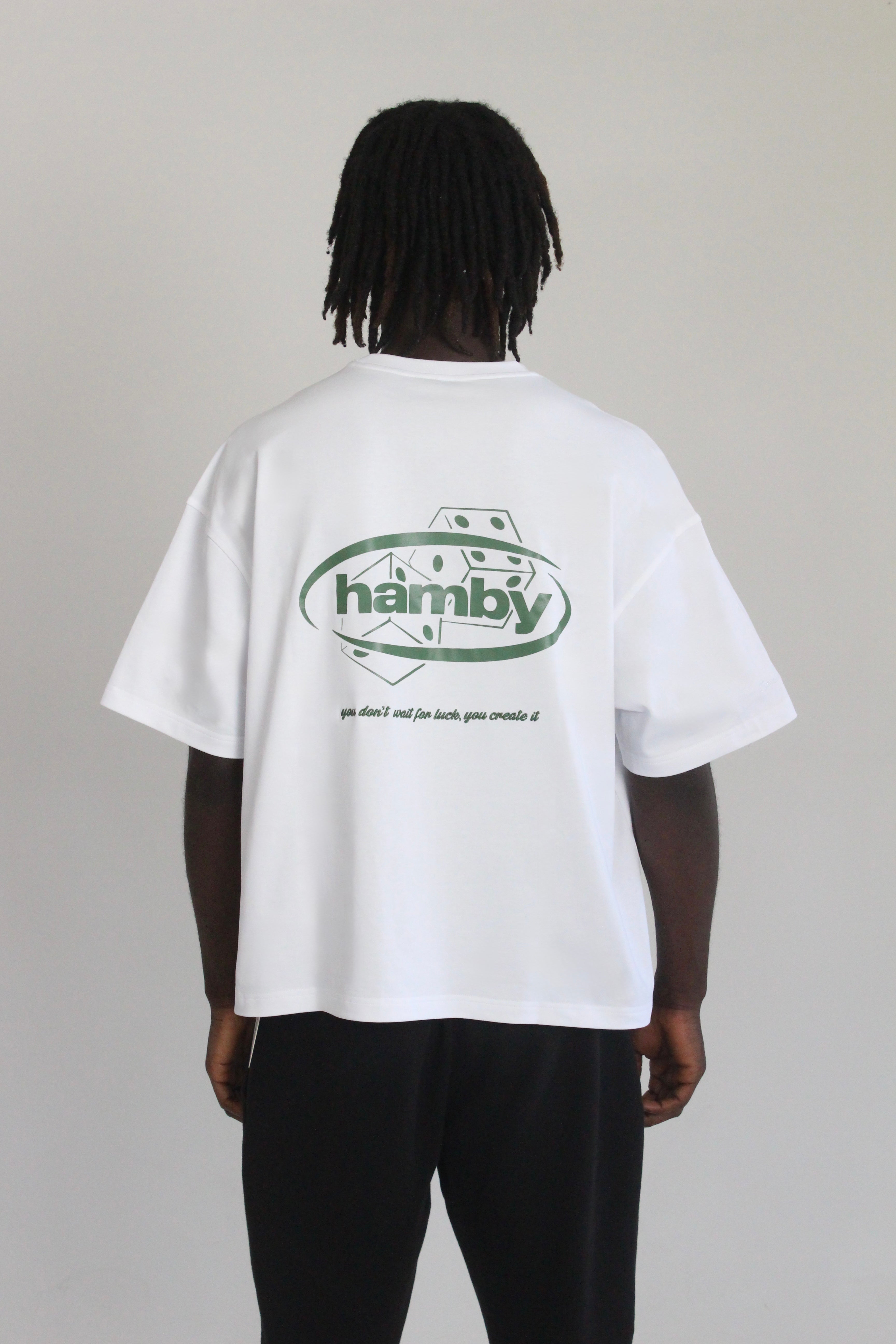 Model wearing white t-shirt with hamby logo and dice screen printed onto back of the tee. Embroidered quote underneath the print. Quote says ‘you don’t wait for luck, you create it’