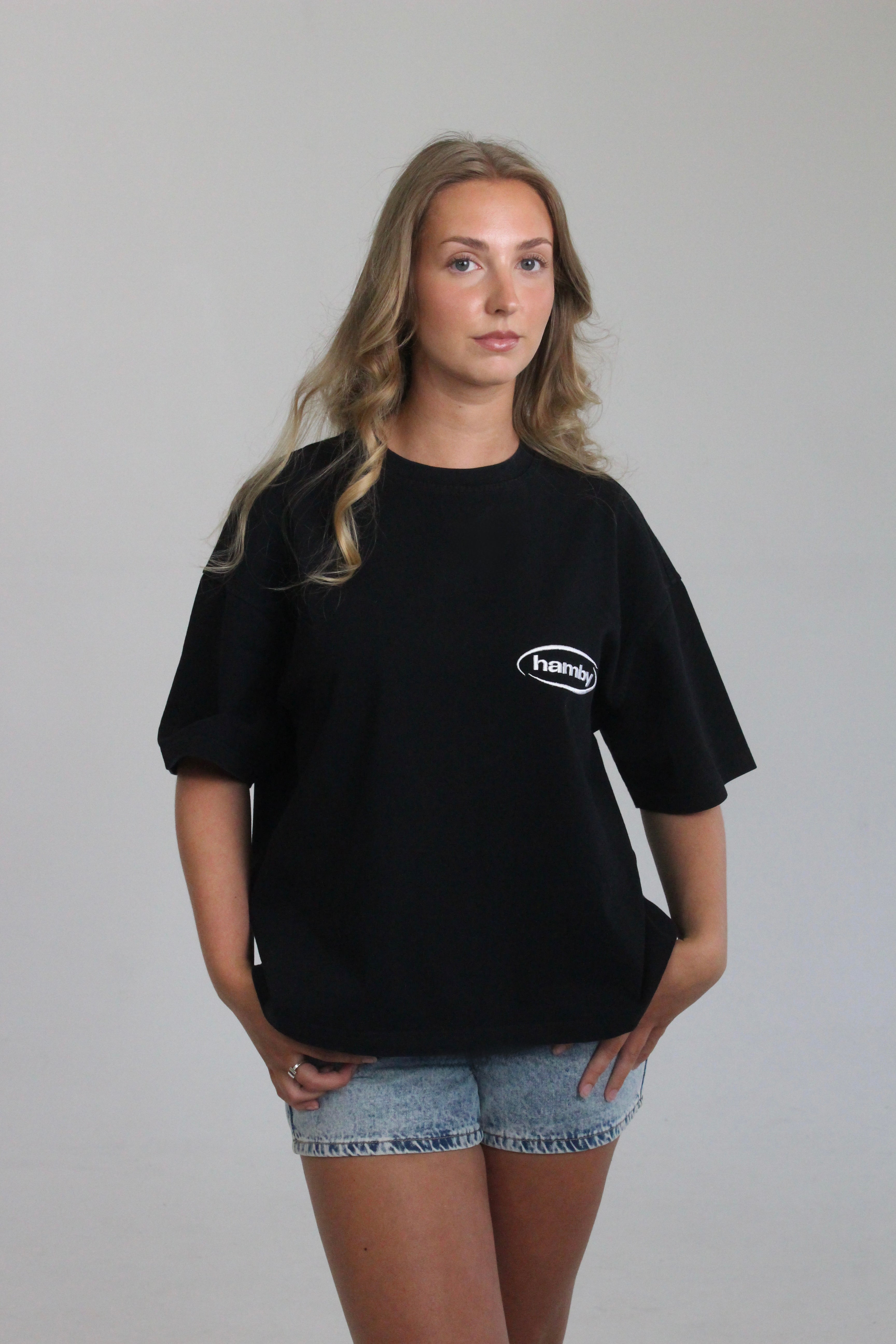 Model wearing boxy oversize hamby black t-shirt with hamby logo embroidered on the front left chest in white. 